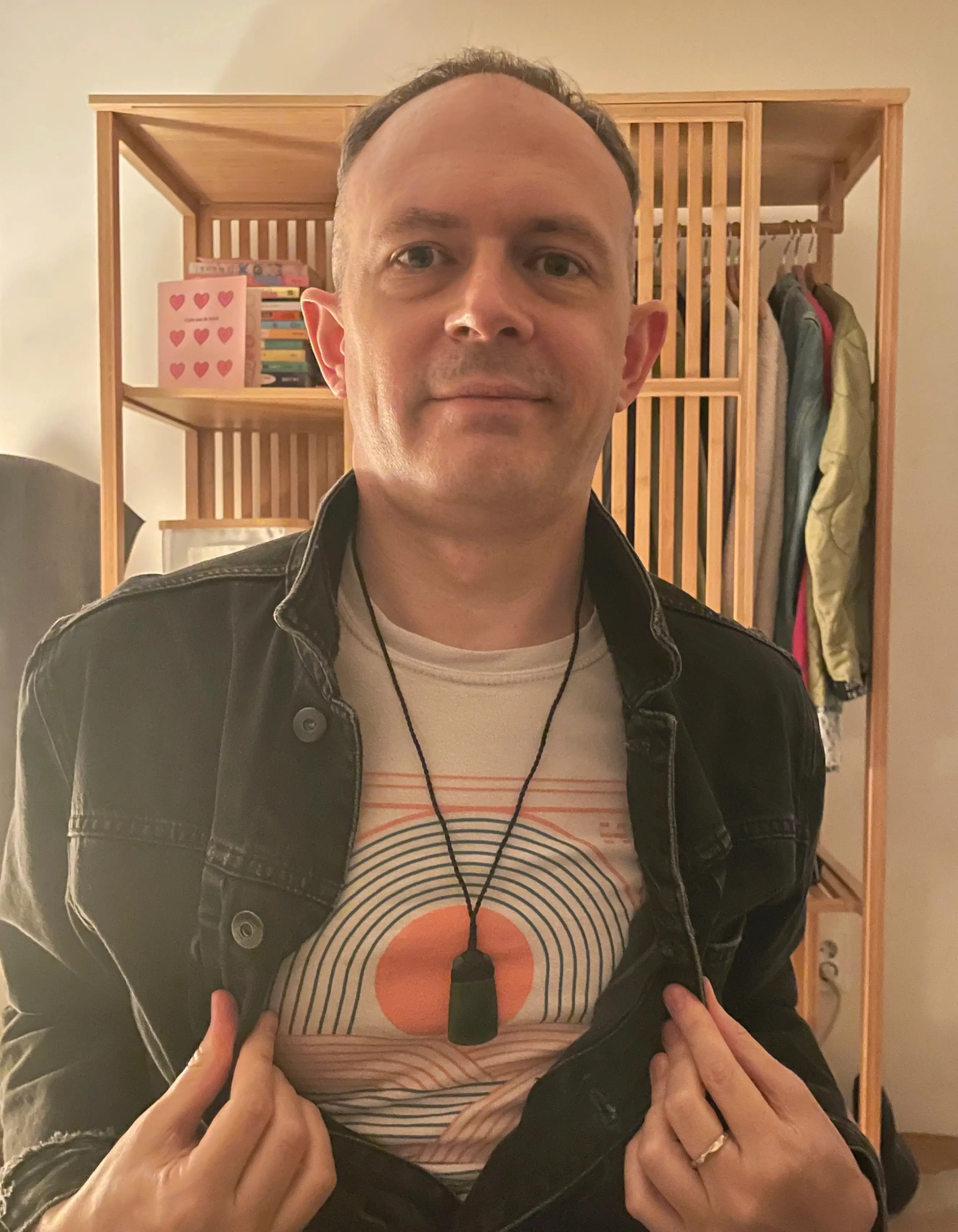 A waist up portrait of a man wearing a black denim jacket. He is holding the jacket open to show his t-shirt and pendant necklace.