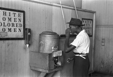 A black man drinking from a water cooler next to a sign that says 'colored.'