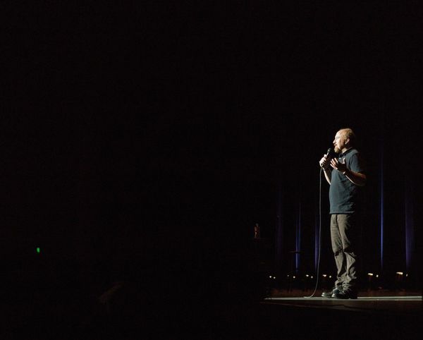 A man standing on stage and speaking into a microphone. He is spotlit and the room is otherwise dark.