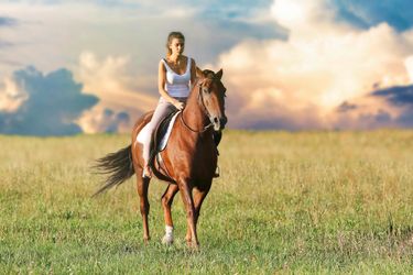 A woman in a singlet riding a brown horse with clouds in the background.