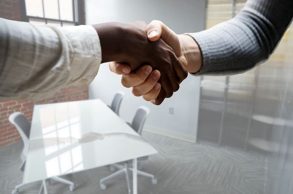 Two hands shaking in an office. There is a table and chairs in the background.