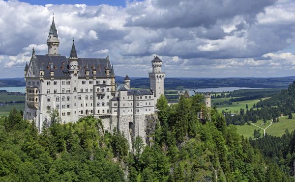 A large stone castle high above the tops of surrounding trees. The castle has several turrets.