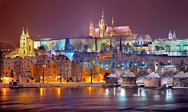 Old buildings in Prague that are lit up at night and seen from across a river.