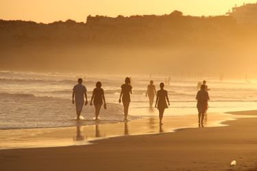 People walking on the beach at the edge of the water. There are cliffs in the background.