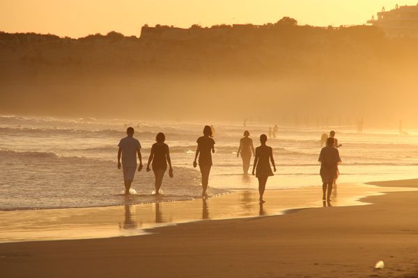 People walking on the beach at the edge of the water. There are cliffs in the background.