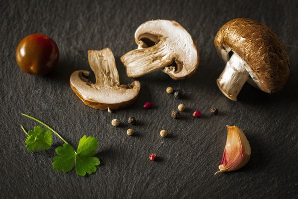 Sliced and whole mushrooms, garlic, peppercorns, and herbs on a dark gray surface.